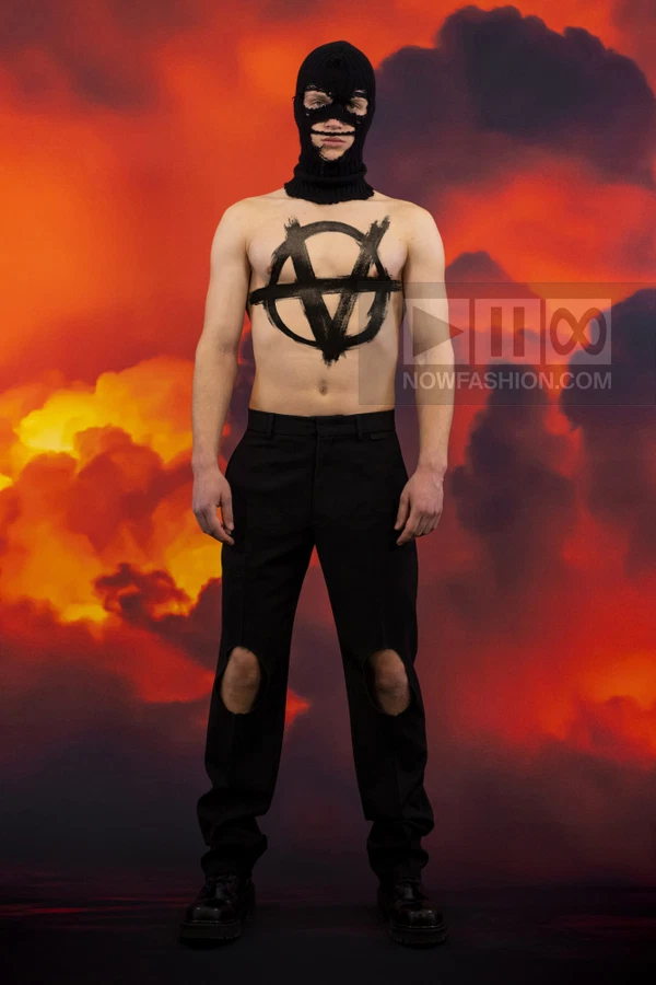 A person wearing a black balaclava and black trousers with knee cut-outs, with a black anarchy symbol painted on their chest.