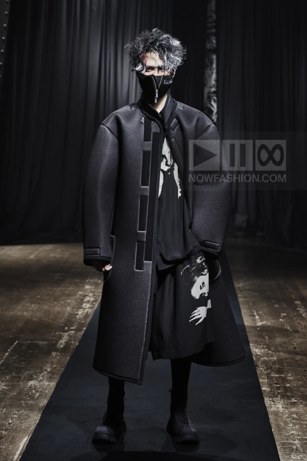 A model on a runway wears a black structured coat with rectangular panel details over a graphic print shirt and shorts.