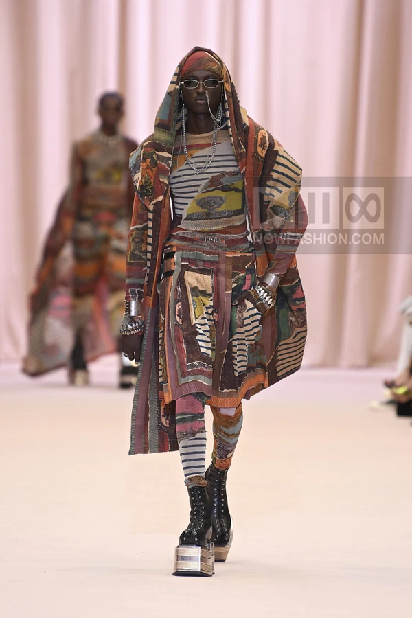 Model wearing a hooded coat, printed top, and skirt over leggings with platform combat boots on a runway.