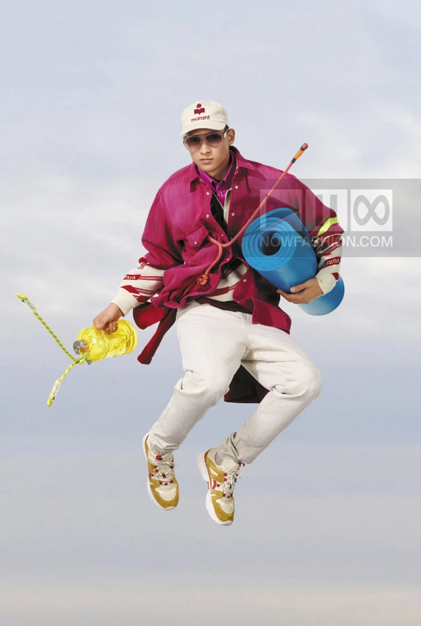 A person jumping in the air wearing a magenta shirt, white pants, a baseball cap, and sneakers, holding a yoga mat.