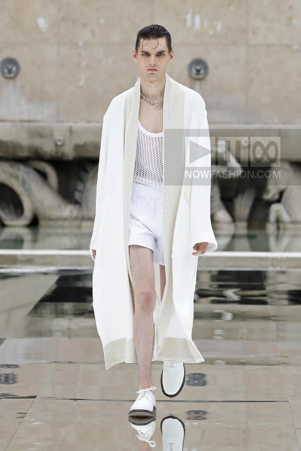 A person walks on a runway wearing a white open-front robe over a white mesh tank top and white shorts.