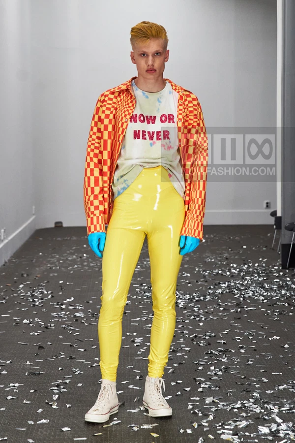 Model wearing a checkered orange shirt, tie-dye slogan t-shirt, and yellow latex pants on a confetti-covered floor.