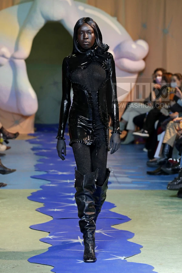Model walking in a black latex mini dress with ruffled trim, sheer glitter leggings, and black leather knee-high boots.
