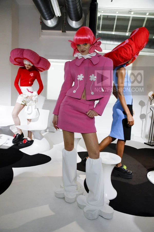 Three models in a studio wearing bright pink, red, and blue outfits with oversized floral-shaped headwear and footwear.