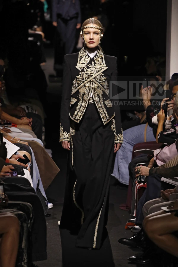 Model wearing a black tailored jacket with gold embroidery and matching wide-leg trousers on a runway.