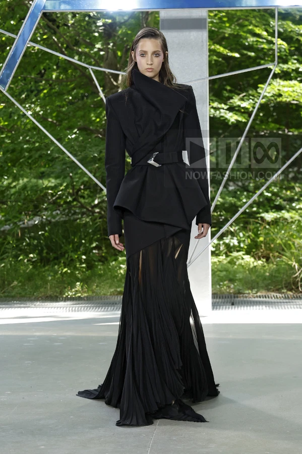 A model wears a black structured jacket with a belted waist and a long, pleated, sheer-panelled skirt on a runway.