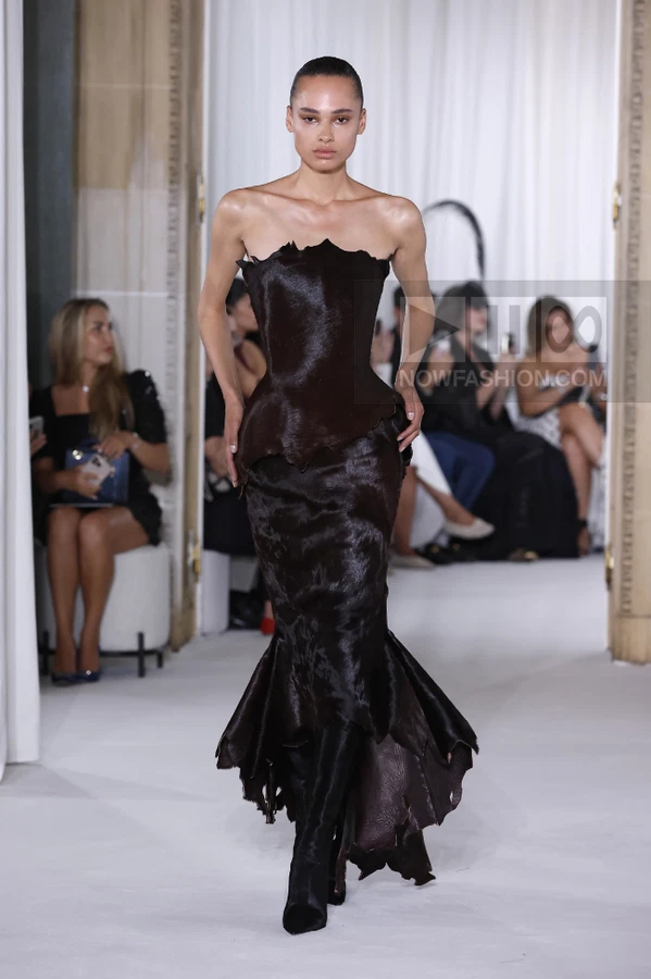 A person walks the runway in a dark brown strapless gown with a textured surface and a flared, uneven hemline.