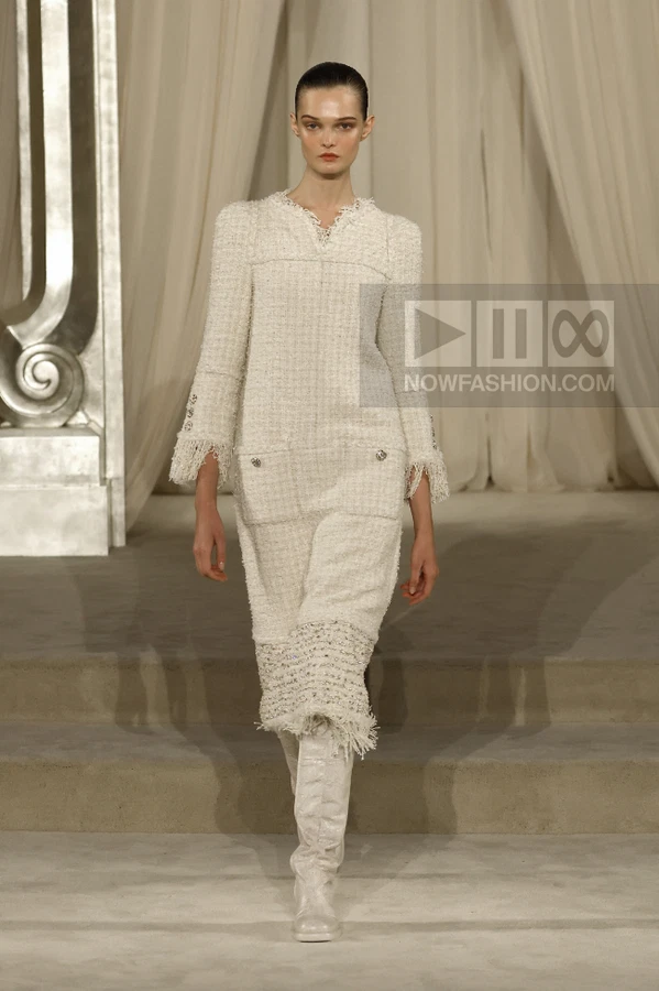 A model walks the runway in a cream-colored tweed shift dress with a fringed hem and matching knee-high boots.