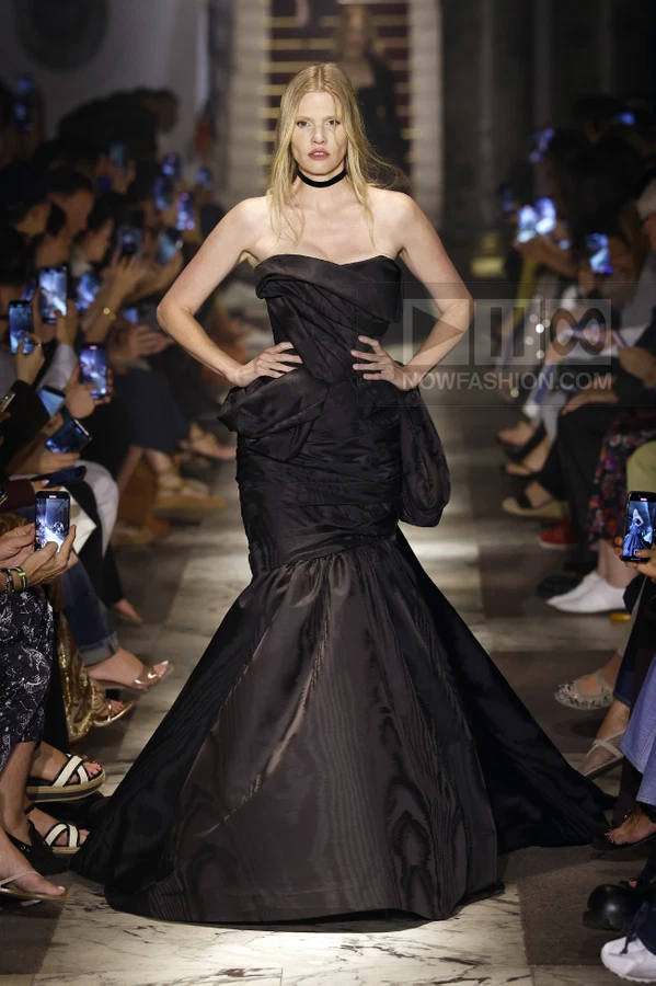 Model wearing a black strapless floor-length gown with a draped bodice and flared skirt on a runway.