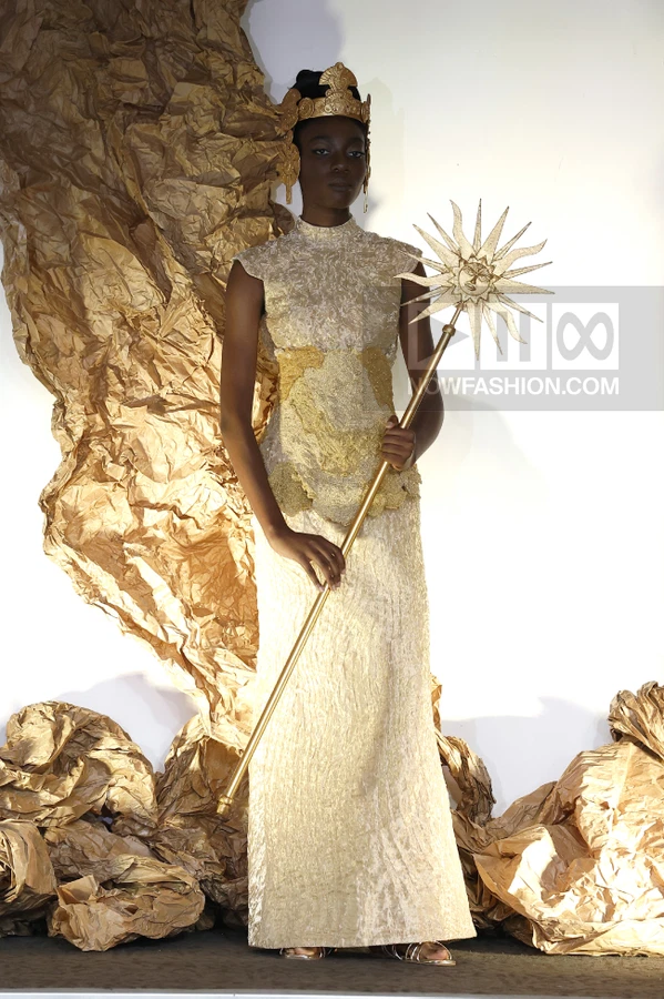 Model wearing a gold textured floor-length gown and crown, holding a sun-shaped staff against a gold paper backdrop.