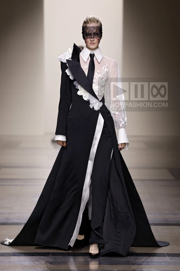 A model on a runway wears a black and white coat-dress with a lace veil, white shirt collar, and floral embroidery.