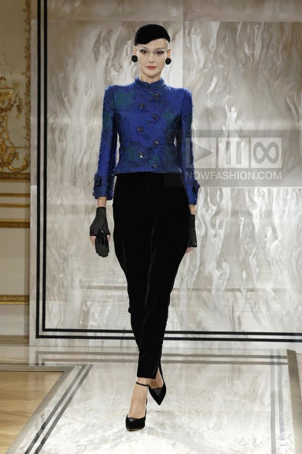A model walks the runway in a blue textured jacket, black velvet trousers, black cap, and black pointed-toe heels.