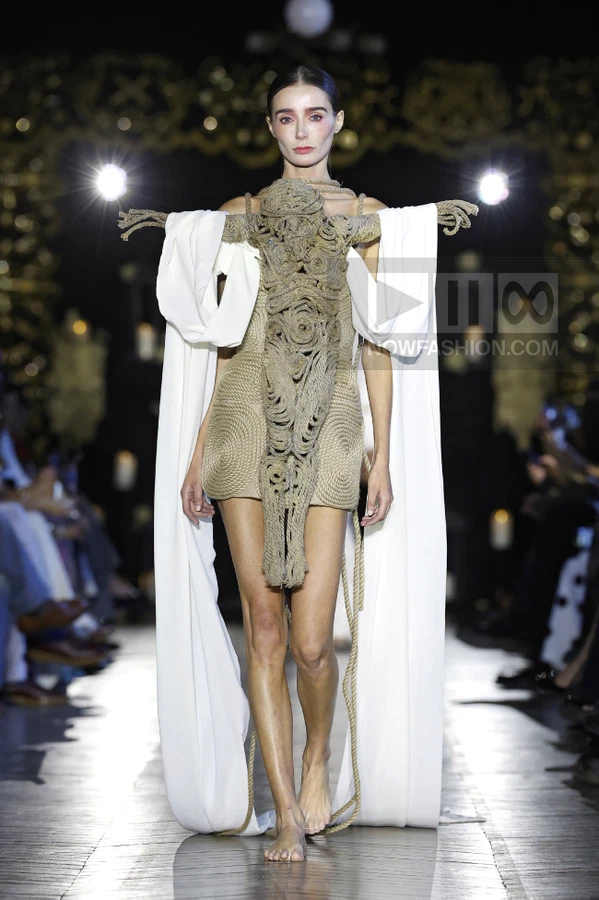 Model walking a runway wearing a beige mini dress with a large, intricate rope-textured chest piece and white cape sleeves.