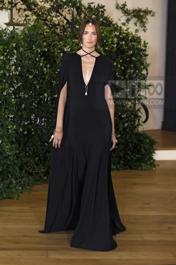 A person wearing a black floor-length gown with a deep V-neckline and cape-style sleeves, standing in front of greenery.