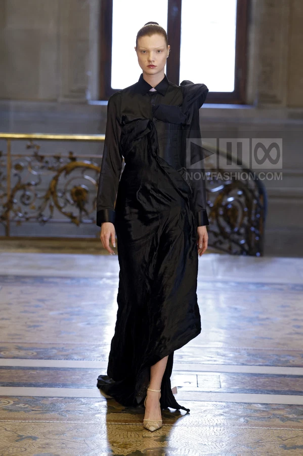 Model wearing a black long-sleeve gown with a draped bodice and asymmetrical hem on a runway.