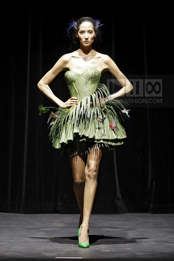Model on runway wearing a green strapless corset dress with a palm-leaf skirt and hummingbird embellishments.