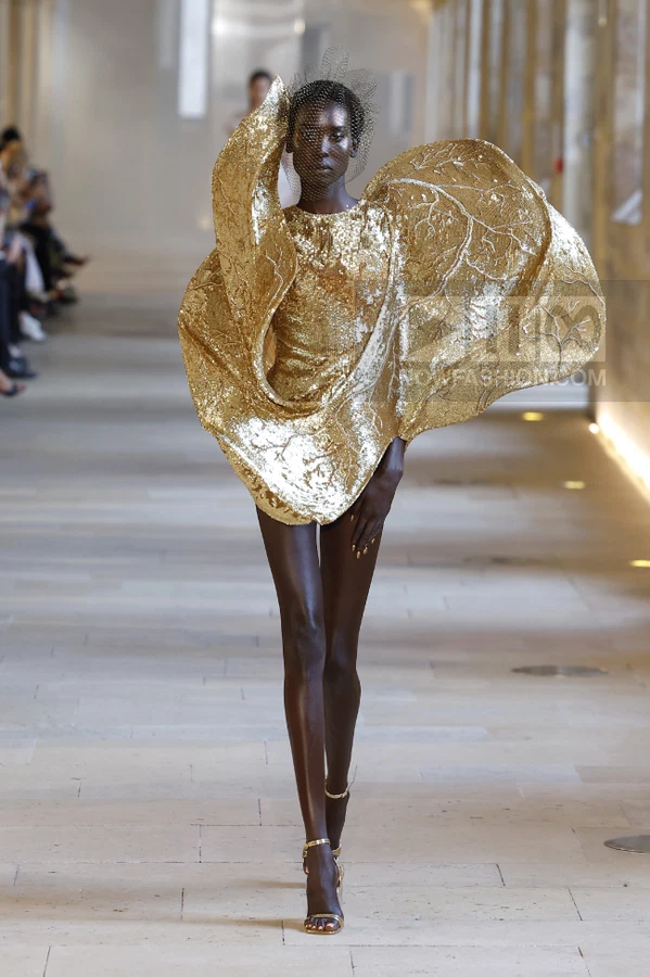 Model walking in a gold sequined mini dress with oversized leaf-shaped structural elements and a black veil.