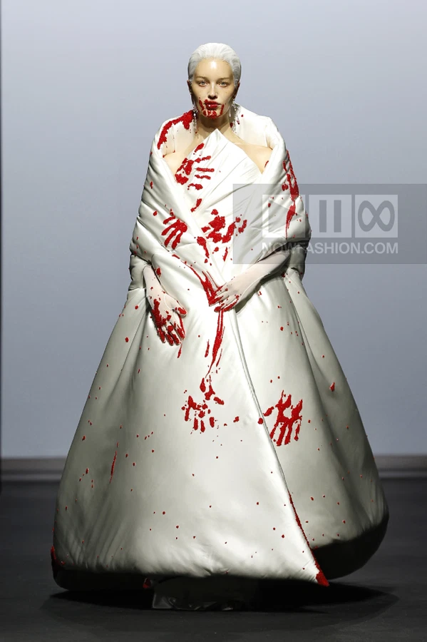 Model wearing a white floor-length quilted gown with red handprint embellishments on a runway.