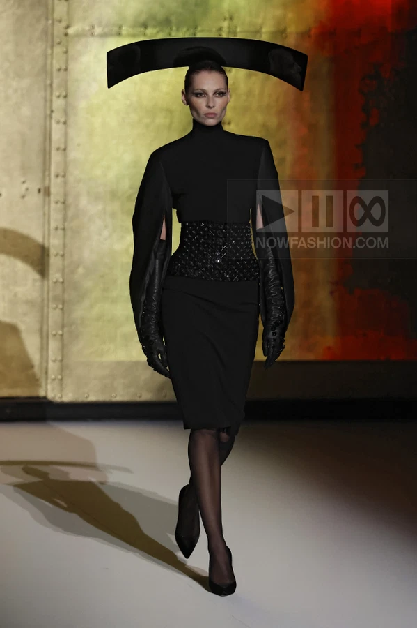 Model wearing a black high-neck dress with a wide embellished waist belt, long gloves, and a large black headpiece.