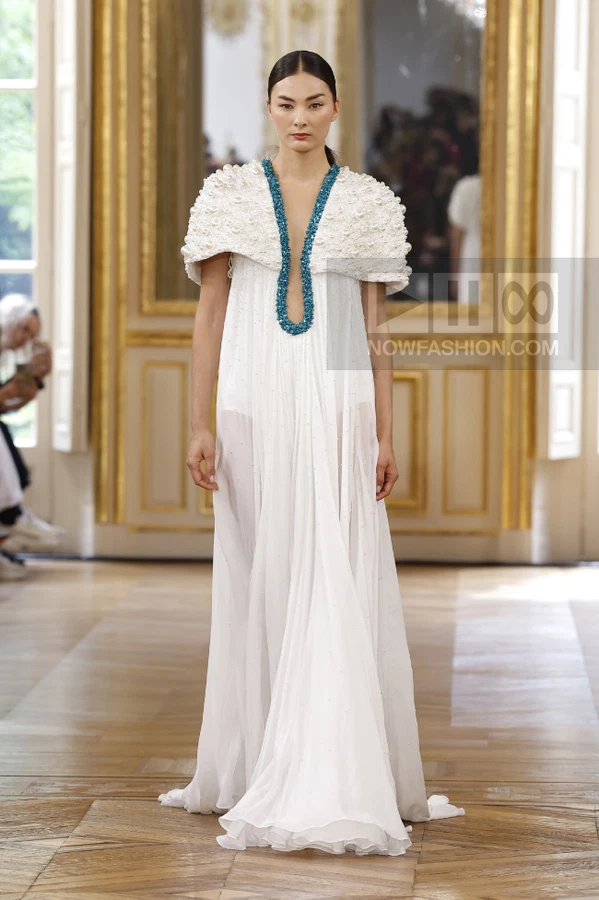 Model wearing a white floor-length gown with a pearl-embellished capelet and a turquoise beaded neckline on a runway.