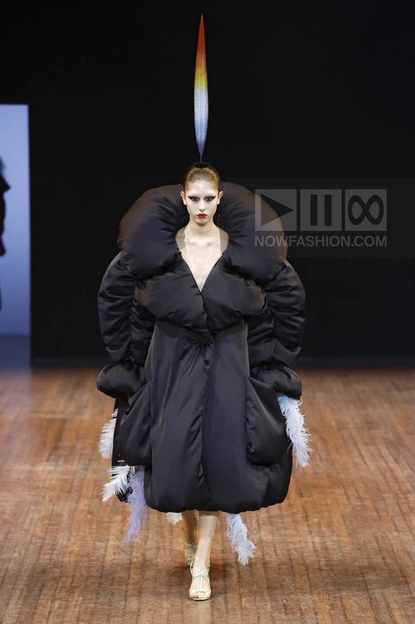 Model wearing a black oversized puffer coat with a large collar and white feather trim, topped with a vertical headpiece.