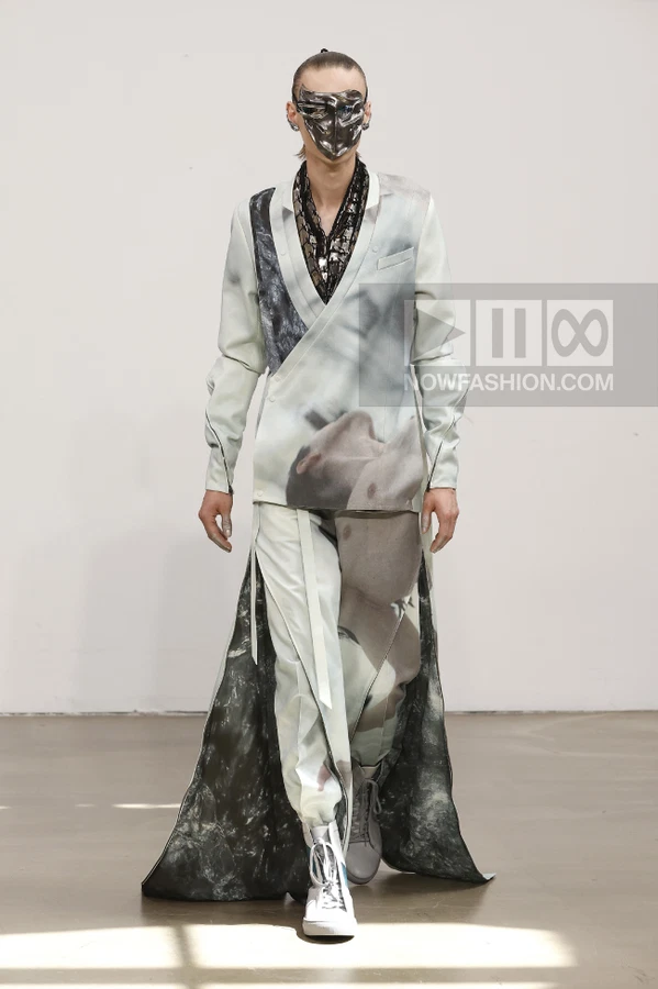 A model on a runway wears a wrap-style jacket and matching trousers with a photographic print and a metallic face mask.