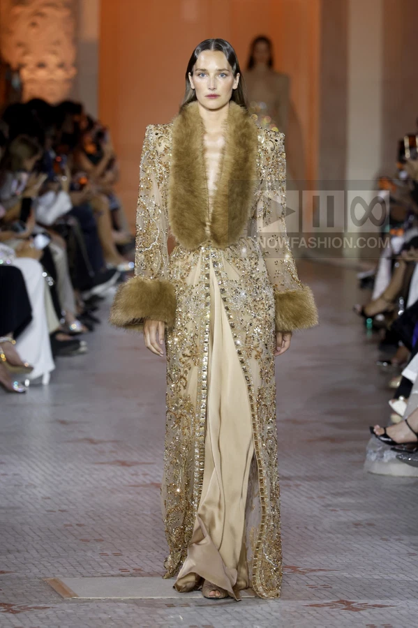 Model walking in a floor-length gold embellished coat with brown fur collar and cuffs over a matching gown.