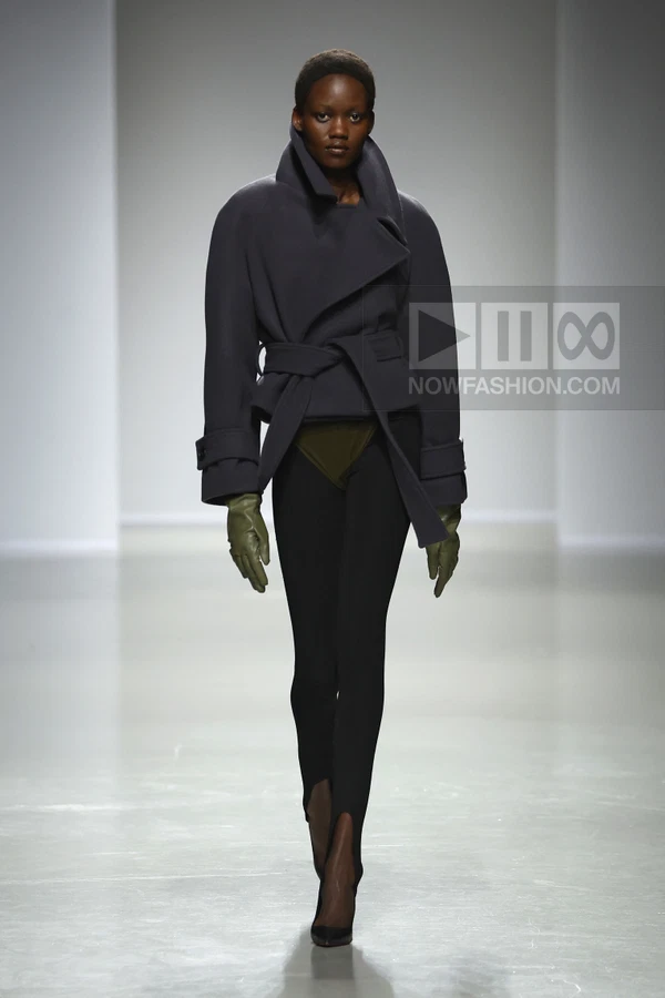 A model walks the runway in a charcoal belted jacket, olive green gloves, and black stirrup leggings.