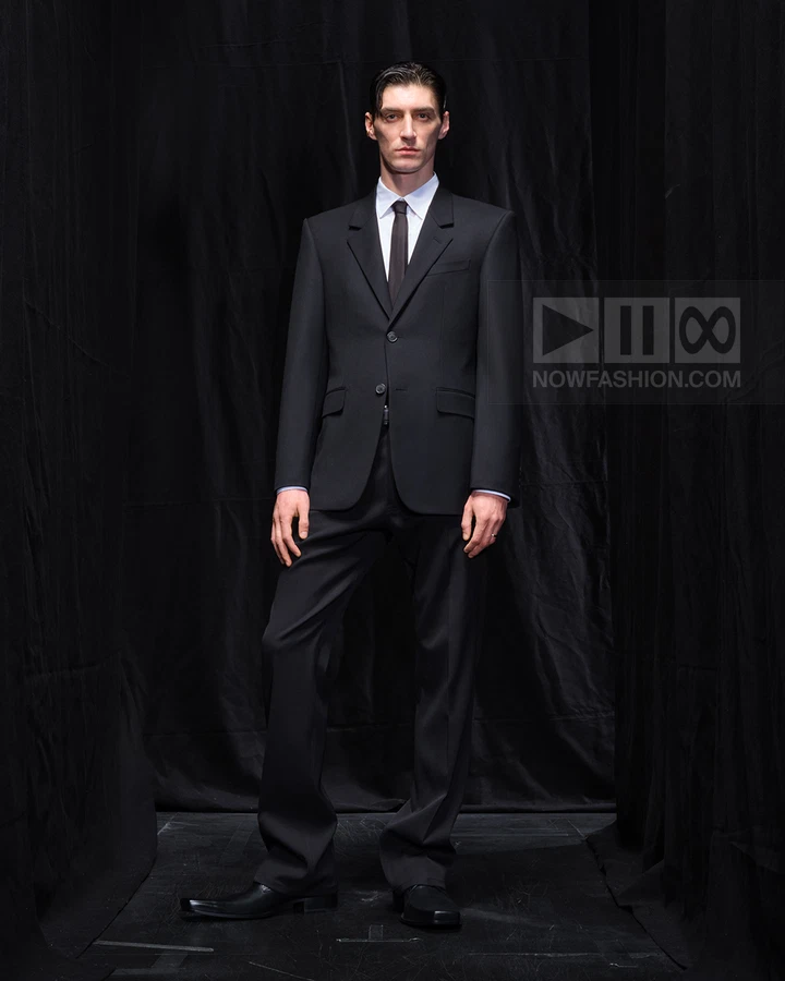 A person wearing a black two-piece suit with a white shirt and black necktie, standing against a black backdrop.