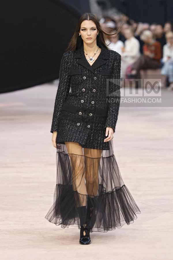 Model walking in a black tweed double-breasted jacket with a sheer tulle skirt overlay on a runway.