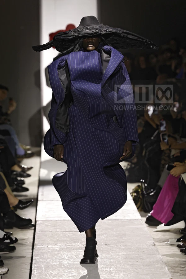 Model wearing a navy pinstripe sculptural coat with a wide-brimmed black hat on a runway.