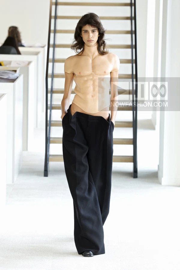 Model wearing a molded anatomical torso piece over wide-leg black trousers on a runway.