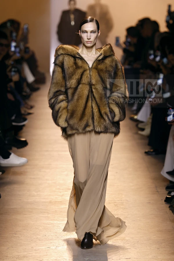 Model walking the runway in a brown fur hooded jacket over a long beige chiffon skirt and black ankle boots.