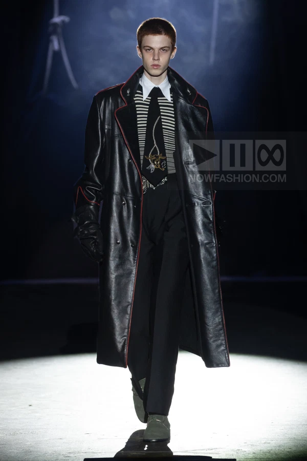 A person walks down a runway wearing a long black leather coat with red piping, a striped shirt, tie, and black trousers.