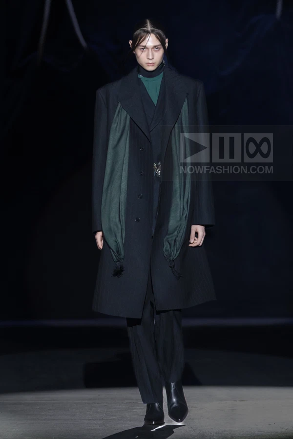 A person walks the runway in a black wool overcoat, dark green scarf, and black trousers against a dark background.