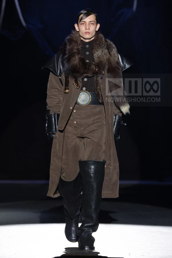 A person walks the runway wearing a brown long coat with a fur collar, black leather gloves, and tall black boots.