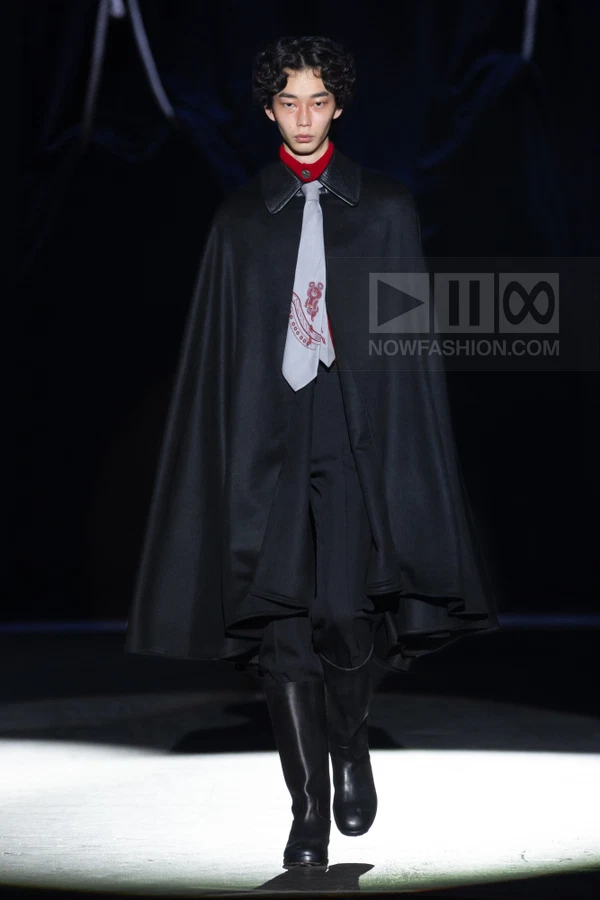 Model wearing a black wool cape over a red shirt and black trousers, with a light gray printed necktie and black boots.