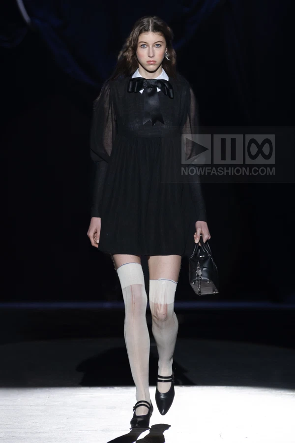 A model walks the runway in a black mini dress with a white collar, black bow, and sheer white stockings with black shoes.
