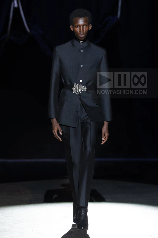 A person wears a black mandarin-collar jacket with a silver-buckled waist belt and matching black trousers on a runway.