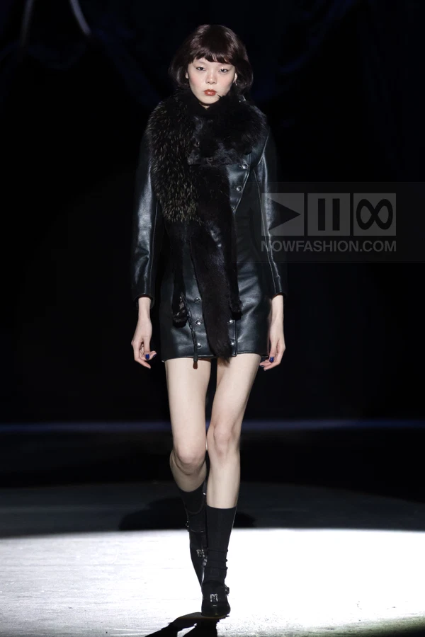 Model walking in a black leather mini dress with a fur stole, black socks, and black buckled shoes on a dark runway.