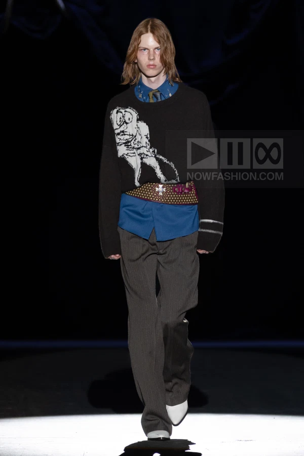 A person walks on a runway wearing a black graphic sweater over a blue shirt, a wide studded belt, and gray trousers.