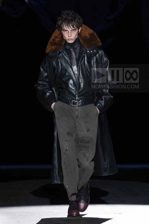 Model wearing a black leather coat with a brown fur collar, dark shirt, tie, and distressed gray trousers on a runway.