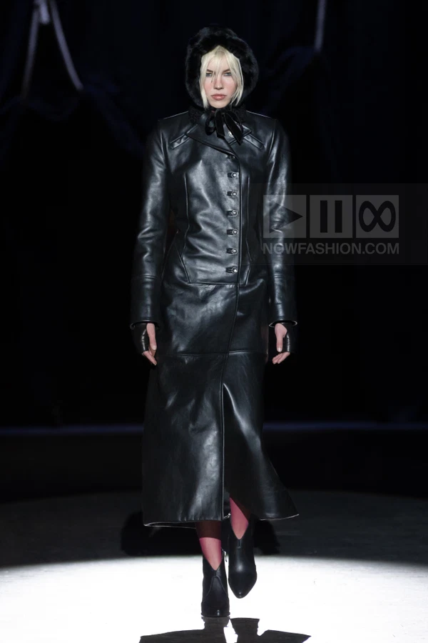 Model in a black leather coat with a fur-lined hood, walking on a dark runway.