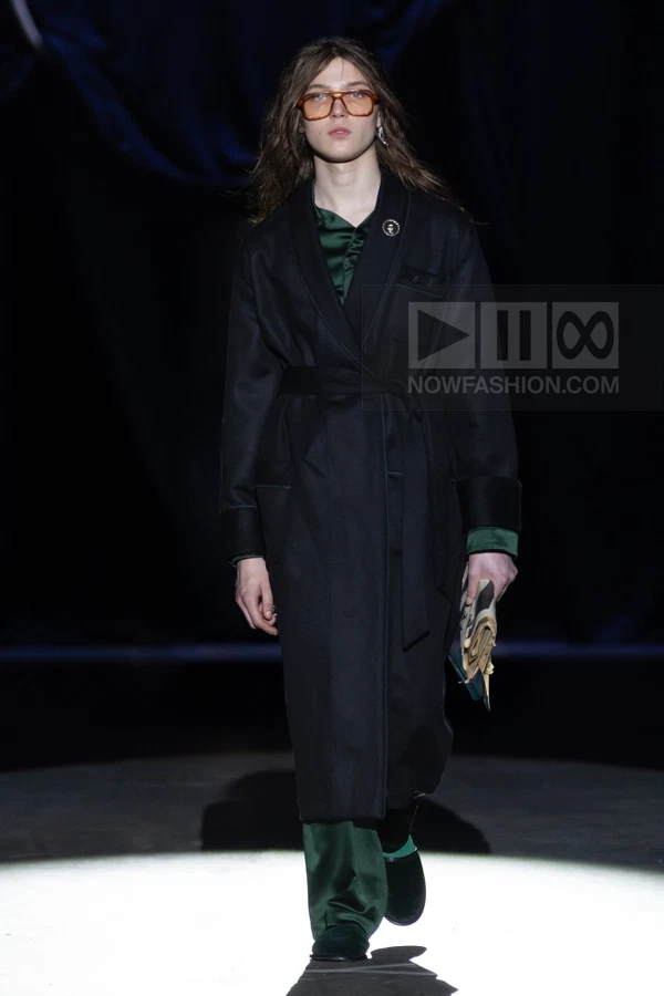 A person walks down a runway wearing a black belted coat over a dark green satin shirt and matching trousers.