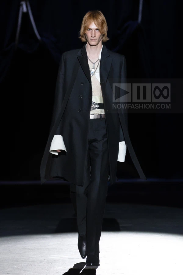 A person walks down a runway wearing a black coat with white cuffs, a white shirt, and black trousers.