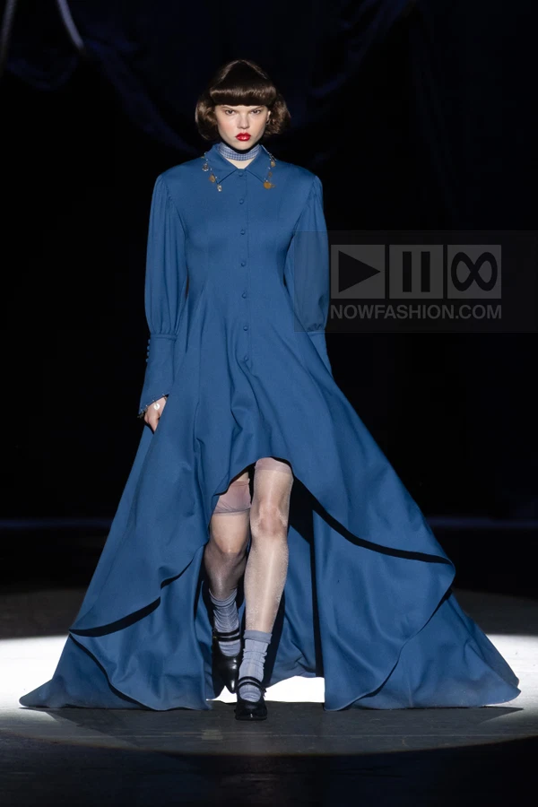Model walks in a blue high-low shirt dress with long sleeves, paired with sheer stockings and black Mary Jane shoes.