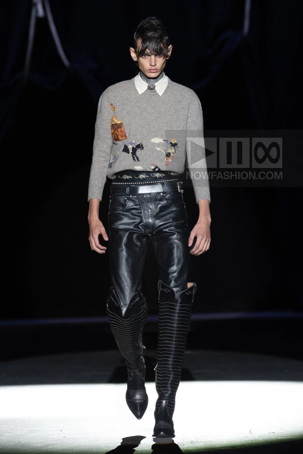 Model walking in a gray embroidered sweater, black leather pants, and tall western-style boots on a dark runway.