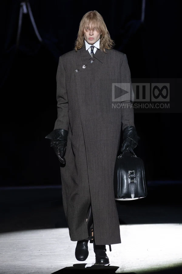 A person walks the runway in a long, dark gray wool coat, white shirt, tie, black leather gloves, and a black holdall bag.