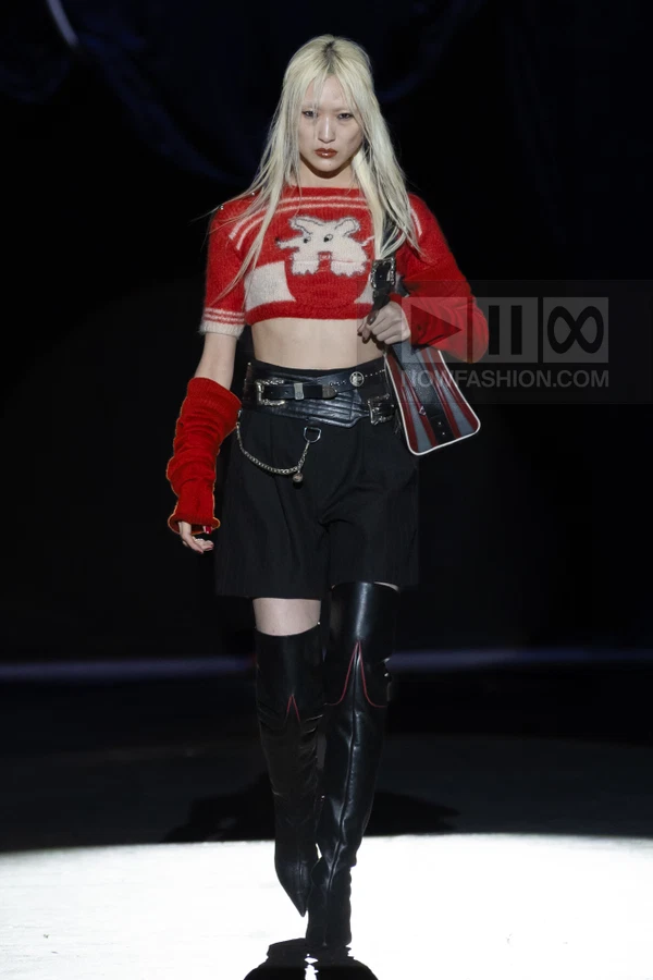 Model walking in a red cropped intarsia sweater, black shorts, leather belt, and over-the-knee boots on a dark runway.