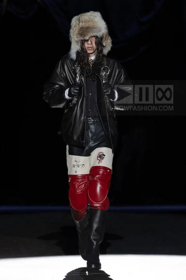 Model wearing a black leather jacket, fur trapper hat, and color-blocked leather pants with red knee panels on a runway.